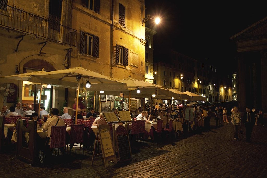 People having dinner at outdoor restaurant tables on an old street at night, under big umbrellas and warm lights
