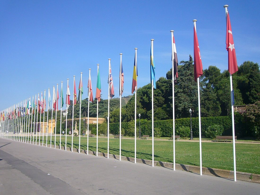 A long row of flags from many countries on tall flagpoles next to a green lawn