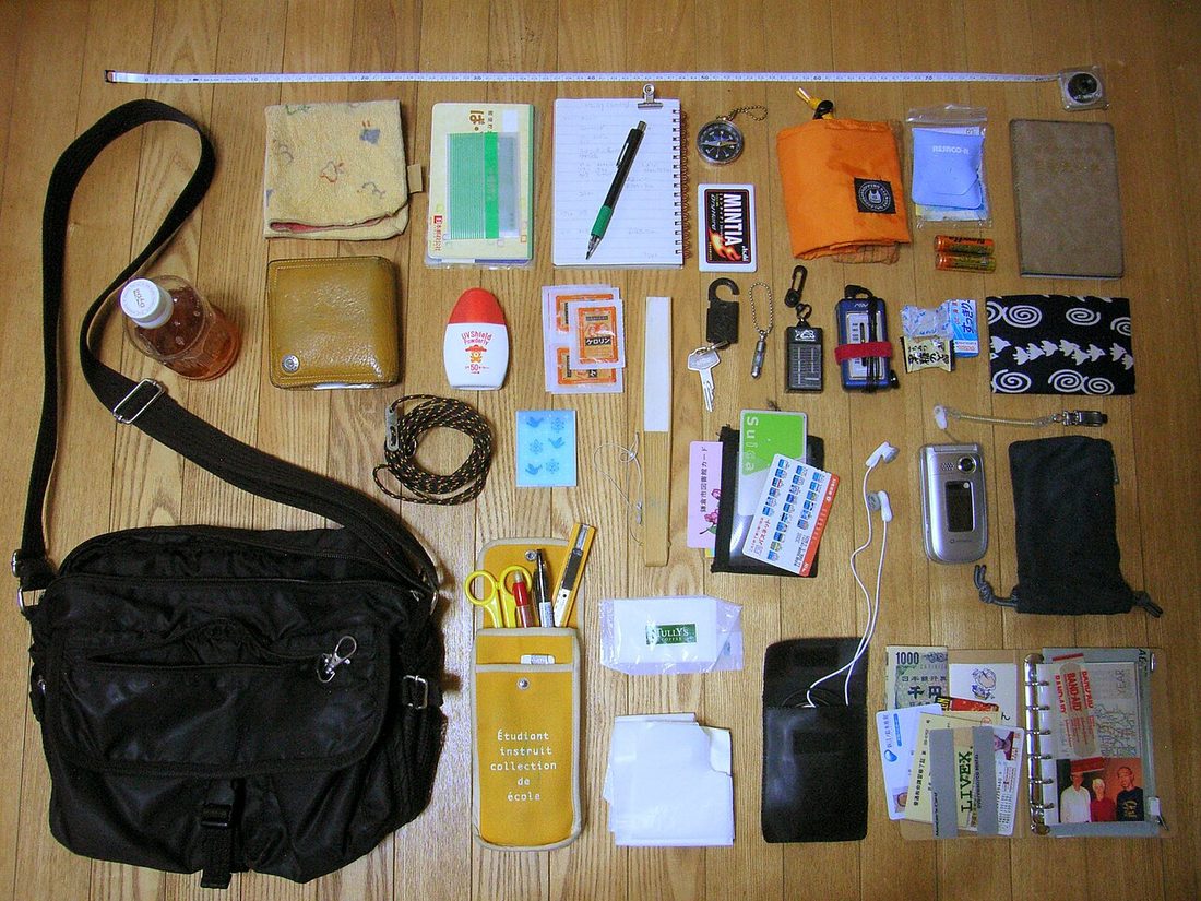 A black shoulder bag and everything from inside it laid out on a wooden floor: pens, cards, a phone, a water bottle, papers and small bags