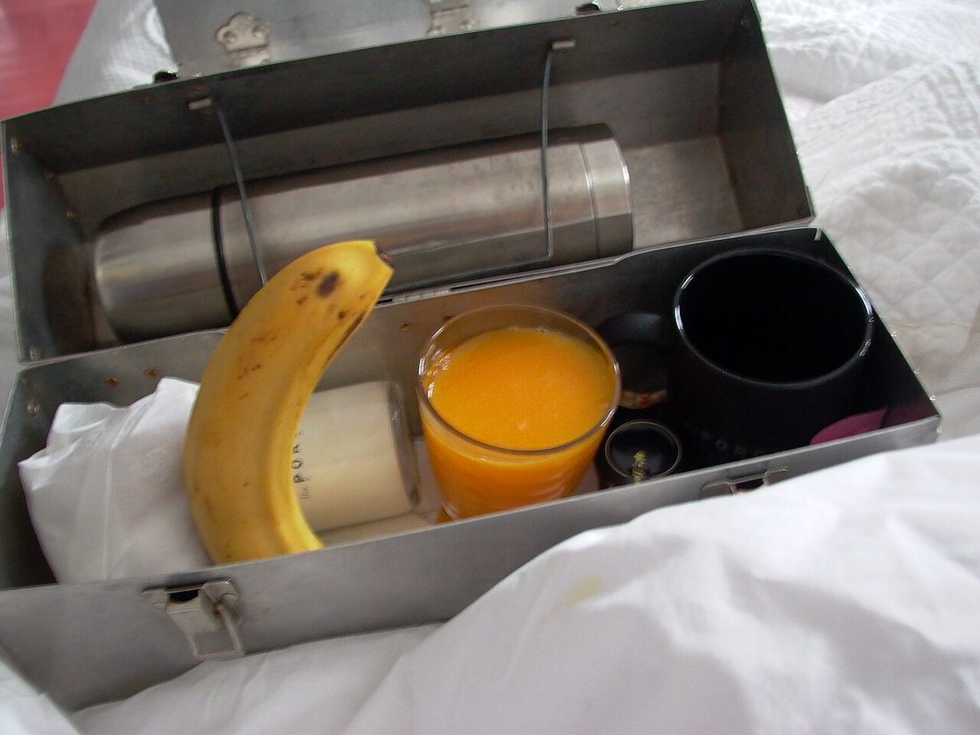 A metal lunchbox with a thermos, a banana, a glass of orange juice and a black cup inside