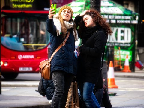Two young women on a city street taking a selfie with a phone, a red bus behind them