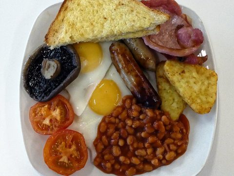 A full English breakfast on a white plate: toast, bacon, two sausages, two fried eggs, a mushroom, tomatoes, baked beans and hash browns