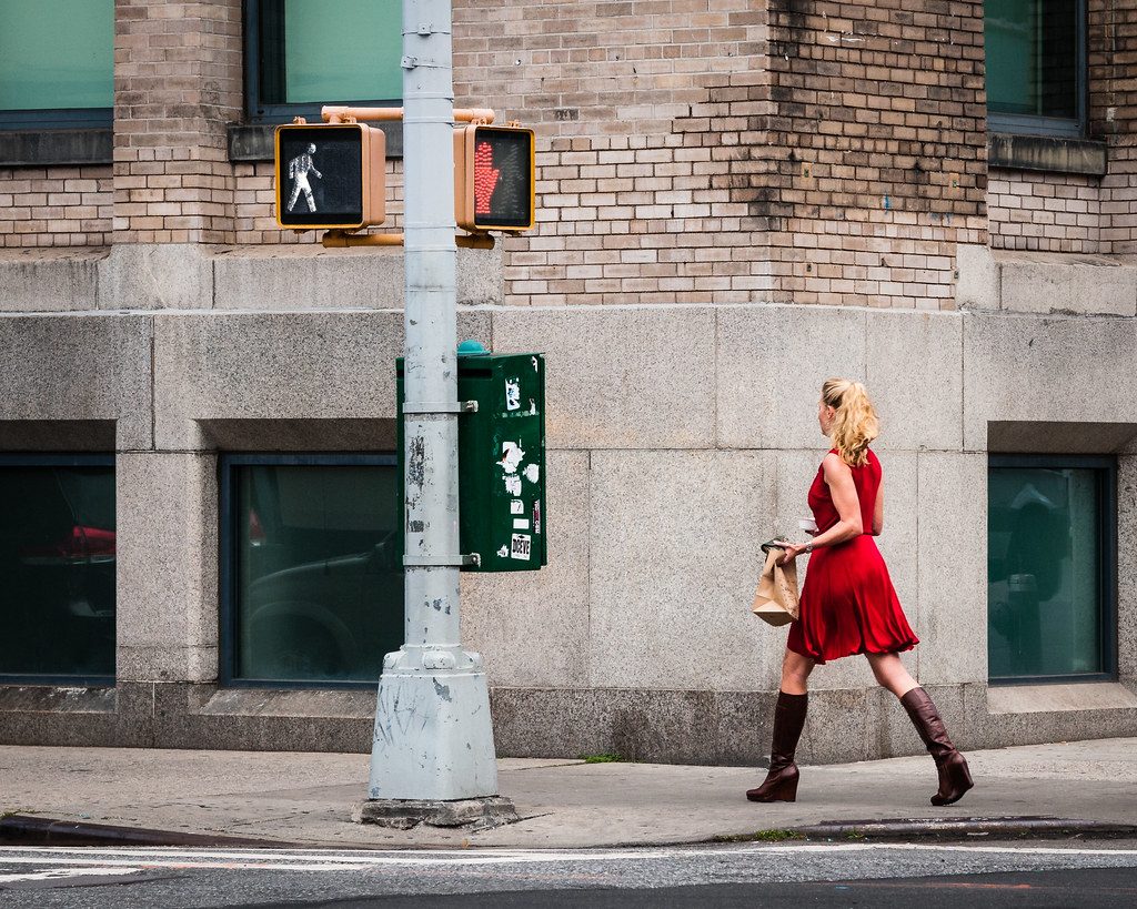 A woman in a red dress and brown boots walking past a crosswalk sign on a New York street