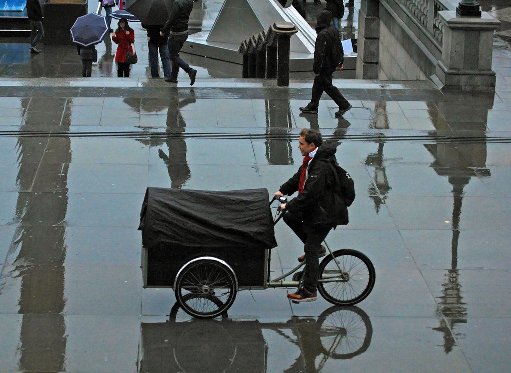 A man riding a cargo bike across a wet square in London, with people holding umbrellas behind him
