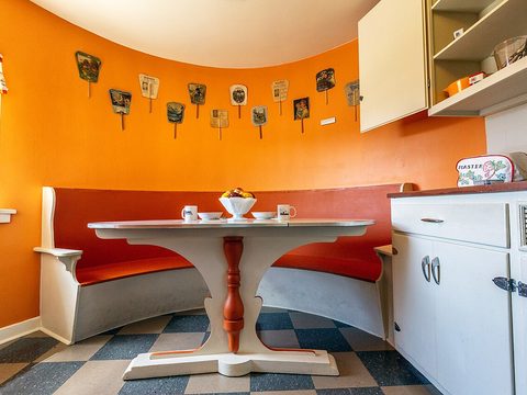 A small kitchen with orange walls, a round table and a curved bench, inside the Shoe House