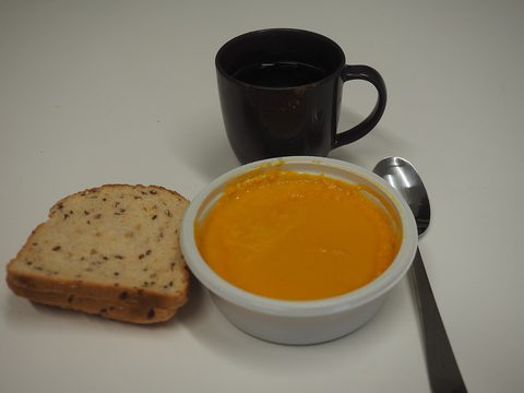 A bowl of orange soup with a slice of bread, a spoon and a black mug