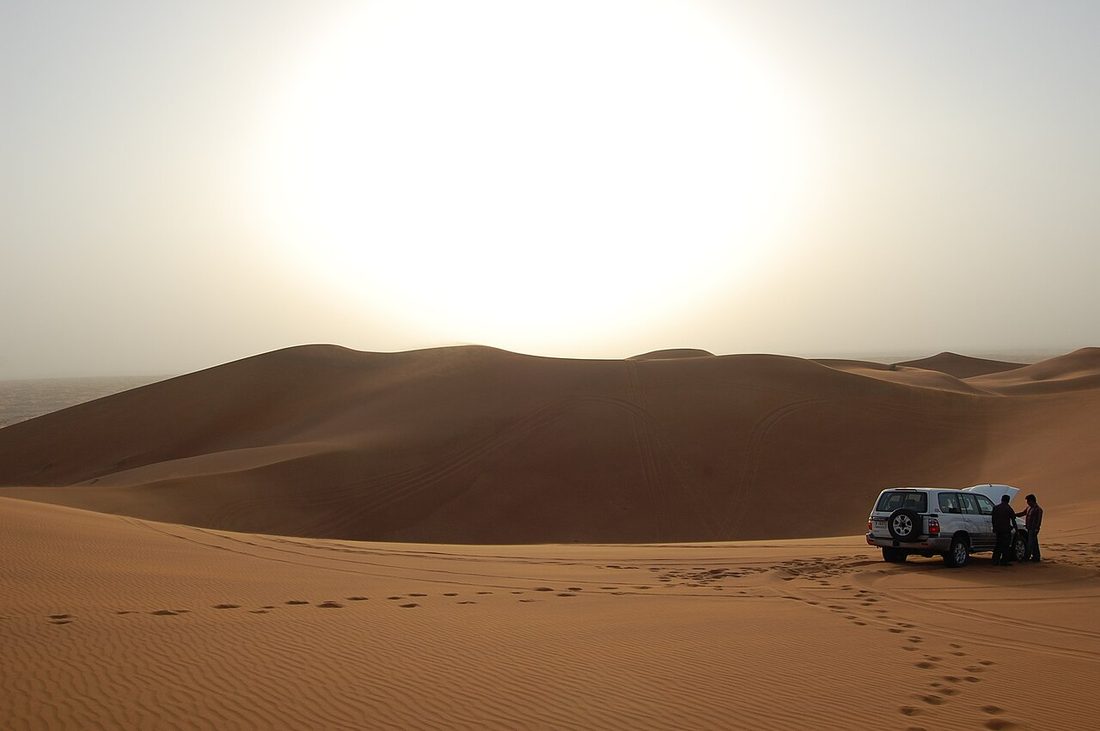 A white SUV on orange sand dunes in the desert near Dubai, with two people next to it and the sun low in the sky