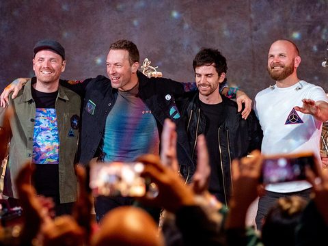 The four members of Coldplay smiling on a stage, with fans holding up their phones in front of them